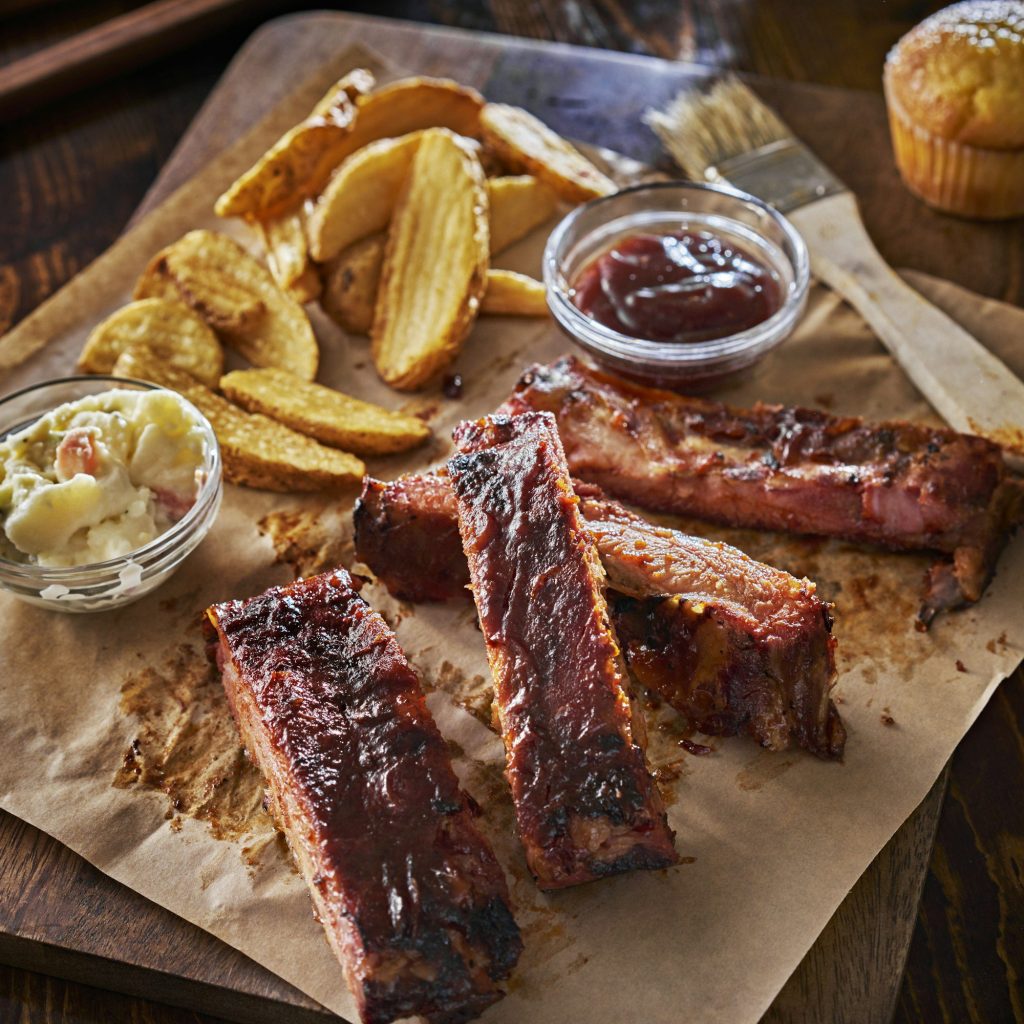 Barbecue ribs with fries, coleslaw, and dipping sauce on a wooden board.